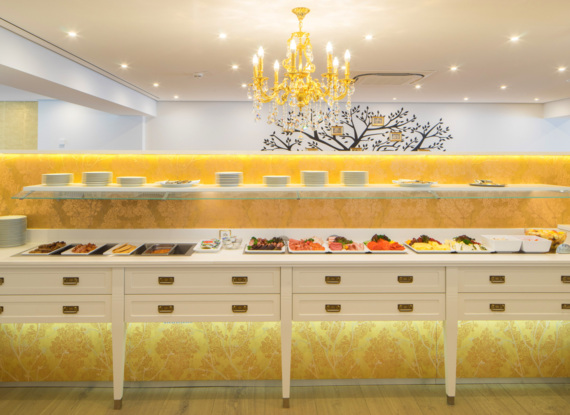 Elegantes Frühstücksbuffet mit verschiedenen Käsesorten, Aufschnitt, Früchten und Tellern unter einem goldenen Kronleuchter in hellem Speiseraum