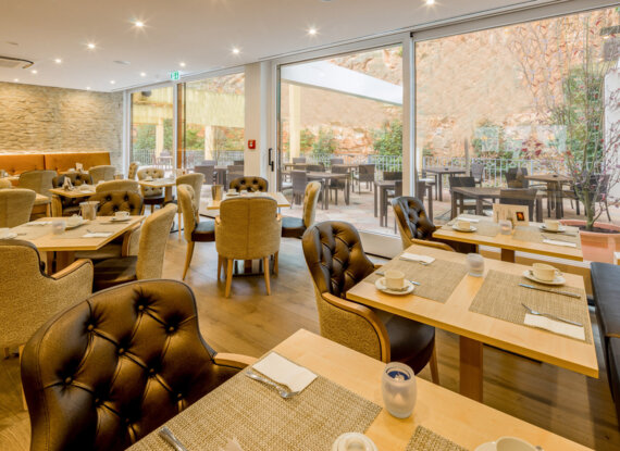Helles Hotelrestaurant mit bequemen Polsterstühlen, Holztischen zum Frühstück und Terrasse mit Sitzgelegenheiten an Steinwand