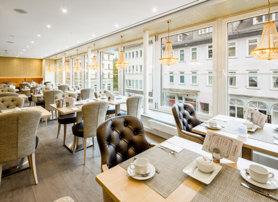 Helles Hotelrestaurant mit großen Fenstern, eleganten Kronleuchtern, gepolsterten Stühlen und liebevoll gedeckten Frühstückstischen.