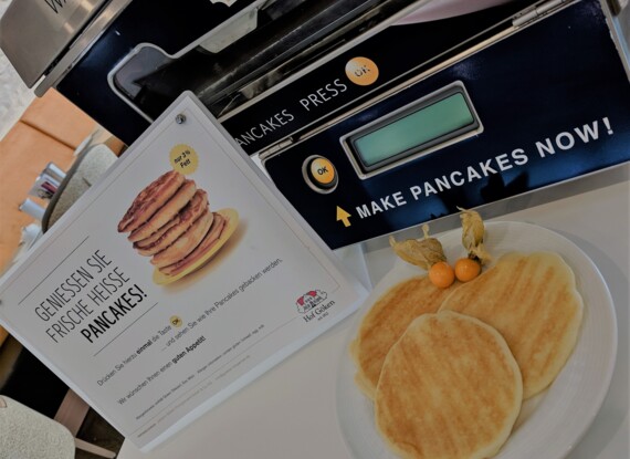 Frisch gebackene Pfannkuchen auf weißem Teller mit Dekobrieseln neben einem Selbstbedienungs-Pfannkuchenautomaten