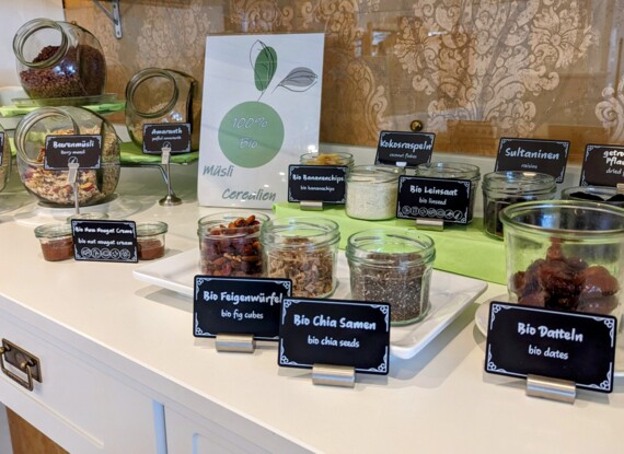 Bio-Frühstücksbuffet mit verschiedenen Cerealien, Trockenfrüchten, Samen und Nuss-Nougat-Creme in Gläsern auf weißer Theke