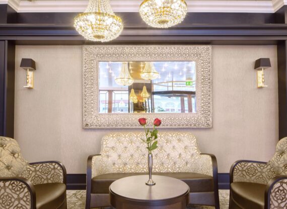 Elegant hotel lounge with patterned armchairs, tufted loveseat, ornate mirror, crystal chandeliers, and vase with two red roses on table