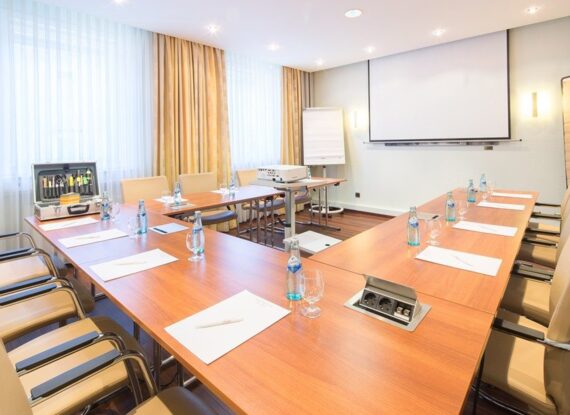 Heller Konferenzraum mit U-förmigen Holztischen, beigen Lederstühlen, Wasserflaschen, Notizblöcken, Beamer und viel Tageslicht