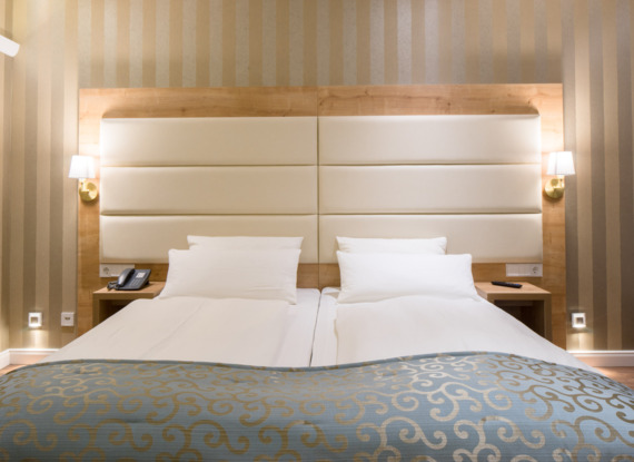 Modernes Hotelzimmer mit zwei Einzelbetten, gepolstertem weißem Kopfteil, beige gestreifter Tapete, Nachttischlampen und blauem Bettüberwurf mit Muster