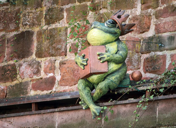 Dekorative Skulptur eines grünen Frosches mit Krone, sitzend auf einem Sims vor einer alten Steinmauer mit Efeu