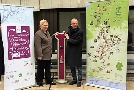Zwei Personen stehen neben einer Ladestation für Elektrofahrzeuge zwischen Informationsbannern zur Deutschen Märchenstraße und Elektromobilität