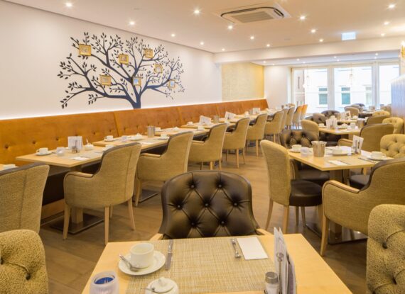Heller Hotel-Speiseraum mit gepolsterten Stühlen, Holztischen zum Frühstück eingedeckt und dekorativem Baum-Wandbild