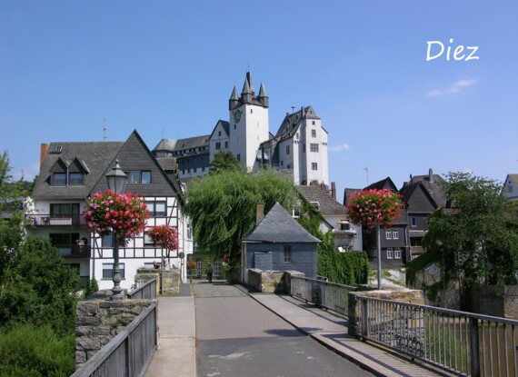 Historische Stadtansicht mit Fachwerkhäusern, blühenden Straßenlaternen und einem Burgturm unter klarem blauem Himmel