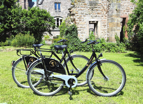 Zwei schwarze Gäste-Fahrräder auf grüner Wiese neben einem historischen Steinbau mit Kletterpflanzen