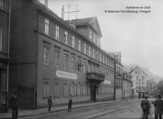 Historische europäische Hotelfassade mit vielen Fenstern, verziertem Balkon und Kopfsteinpflasterstraße mit Fußgängern um 1920