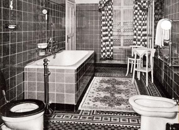 Vintage-Badezimmer im Hotel mit gefliesten Wänden und Boden, Badewanne, Bidet, Toilette, gemustertem Teppich und Tisch mit zwei Stühlen am Fenster mit Vorhängen
