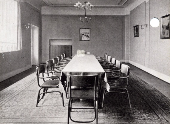 Heller Konferenzraum mit langem, gedecktem Tisch, umgeben von 14 modernen Armlehnstühlen und schlichter Einrichtung