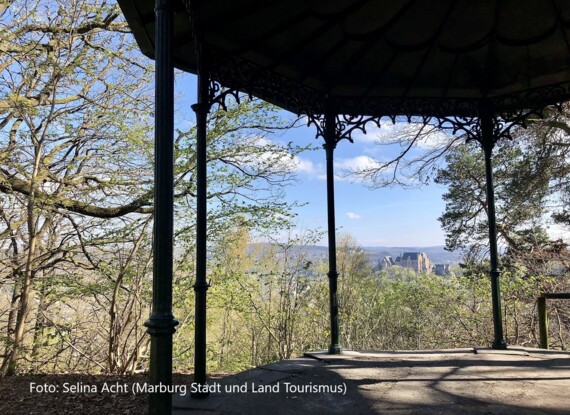 Verzierte eiserne Pergola mit Blick auf grüne Bäume und eine ferne historische Burg bei blauem Himmel