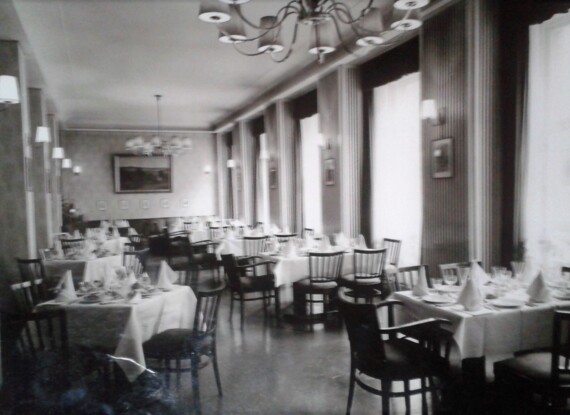 Klassischer Speisesaal mit weißgedeckten Tischen, Holzstühlen, Kronleuchtern und großen Fenstern mit Vorhängen