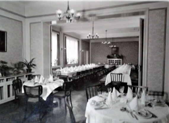 Eleganter Speisesaal mit langen Tischen, weißem Leinen, gefalteten Servietten, Gläsern und Holzstühlen unter Kronleuchtern