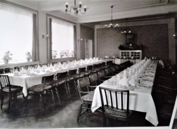 Eleganter Speisesaal mit langen Tischen, weißer Tischwäsche, gefalteten Servietten, Gläsern und Stühlen, bereit für eine große Veranstaltung.