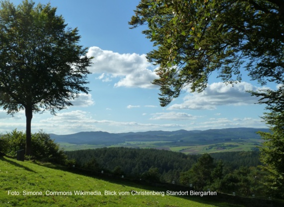 Blick von einem Hügel mit Bäumen im Vordergrund auf grüne Felder und bewaldete Hügel unter blauem Himmel mit Wolken
