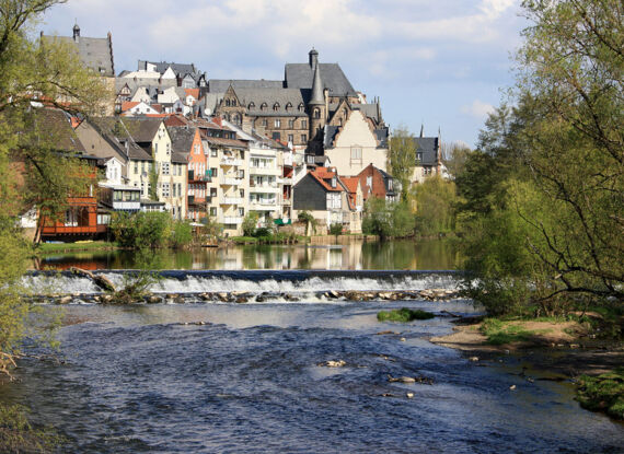 Charmante Stadt am Fluss mit historischer Architektur, geneigten Dächern und grünen Bäumen entlang eines ruhigen Flusses unter blauem Himmel