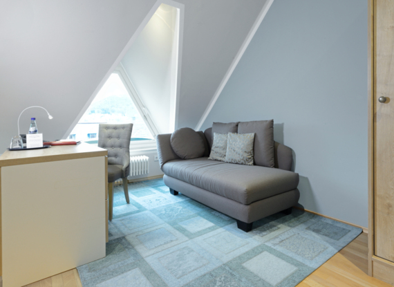 Gemütliche Hotelsitzecke mit grauem Chaiselongue, gemusterten Kissen, Schreibtisch und dreieckigem Fenster für Tageslicht.