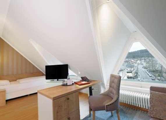 Gemütliches Mansarden-Zimmer mit Einzelbett, Holztisch, gepolstertem Stuhl, Fernseher und dreieckigem Fenster mit Blick auf Stadt und Hügel