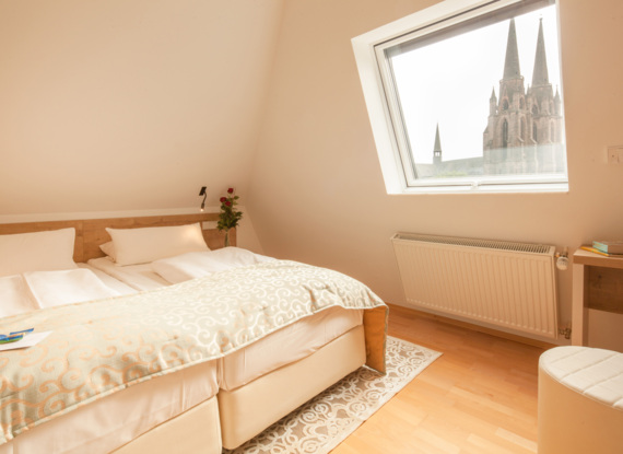 Helles Hotelzimmer mit zwei Einzelbetten, gemustertem beigen Bettüberwurf, Holzboden und Dachfenster mit Blick auf Kathedraltürme