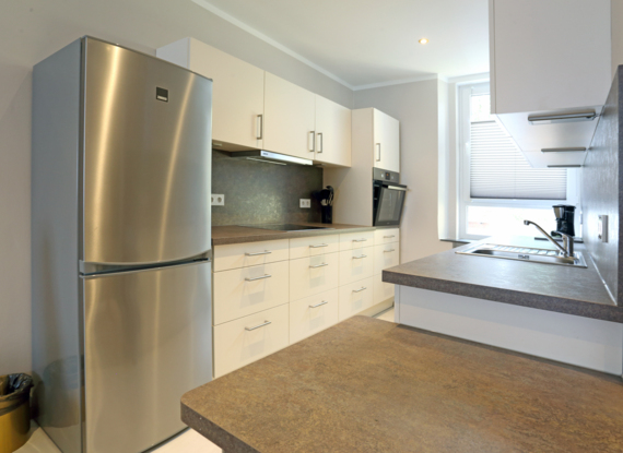 Moderne Hotelküche mit Edelstahlkühlschrank, eingebautem Ofen, großzügiger Arbeitsfläche und hellem Fenster mit Jalousien