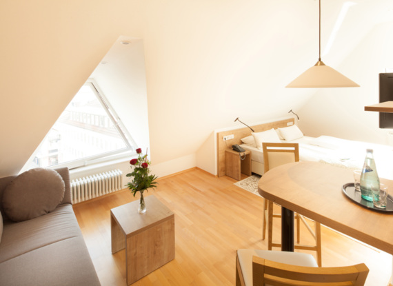 Helles Dachzimmer mit schräger Decke, Holzboden, Doppelbett, Sofa, kleinem Tisch mit Blumen und Essbereich mit Pendelleuchte