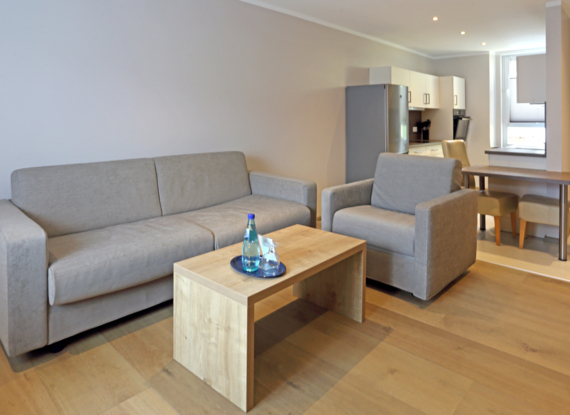 Modernes Suite-Wohnzimmer mit beigem Sofa und Sessel, Holztisch mit Wasserflaschen, Kochnische und Esstisch mit Stühlen.