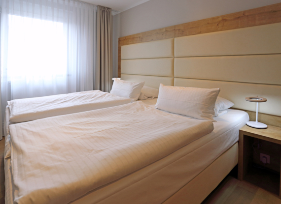 Helles Hotelzimmer mit zwei Einzelbetten, gepolstertem beigem Kopfteil, weißer Bettwäsche, Holznachttisch und sanftem Tageslicht