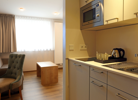 Kleine Hotel-Suiten-Küche mit Mikrowelle, Elektroherd, Spüle, Wasserkocher und gemütlichem Sitzbereich mit Sessel und Holztisch
