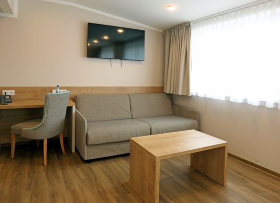 Helles Hotelzimmer mit Holzboden, beigem Sofa, Holztisch, Schreibtisch mit Stuhl und wandmontiertem Flachbildfernseher