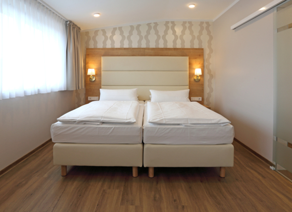 Modernes Zweibettzimmer mit zwei Einzelbetten, gepolstertem Kopfteil, warmen Holzakzenten, gemustertem Tapeten und Tageslicht vom großen Fenster