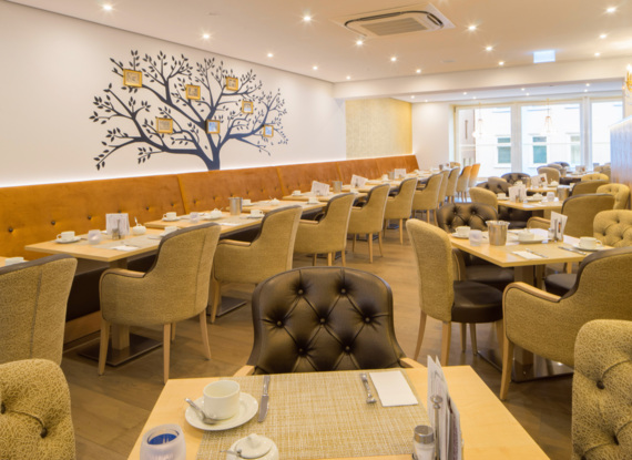 Heller Hoteldiningbereich mit gesteppter Polsterung, senffarbenem Bankett, Wanddekor in Form eines Baums und gedeckten Frühstückstischen.