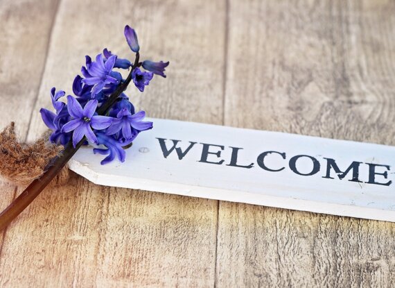 Weißes Holzschild mit dem Schriftzug 'Willkommen' und leuchtend violetten Blumen auf natürlicher Holzoberfläche