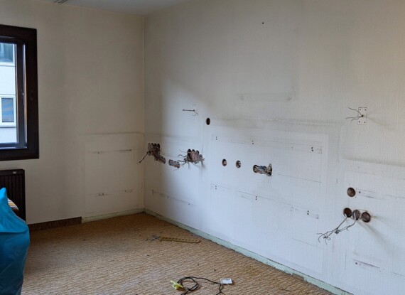 Empty hotel room under renovation with exposed wiring on textured white walls and natural light from a large window