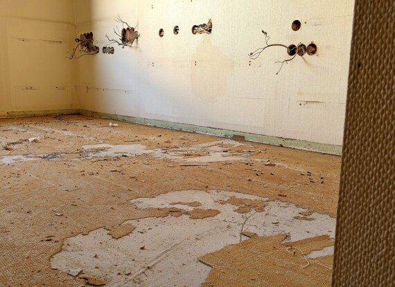 Empty room under renovation with stripped flooring and exposed wiring protruding from beige walls