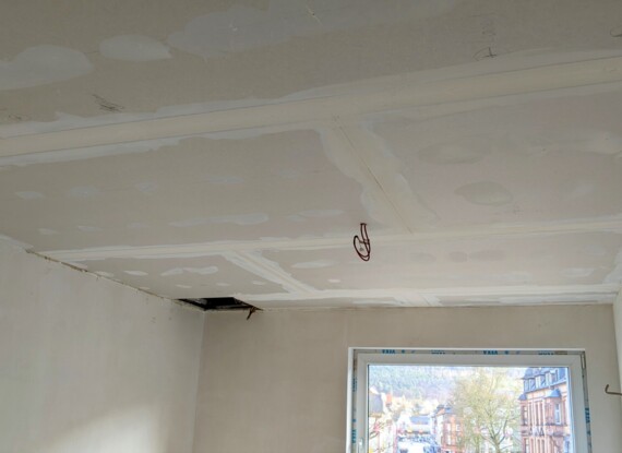Room under renovation with unfinished drywall ceiling and exposed wiring, window showing urban street view outside