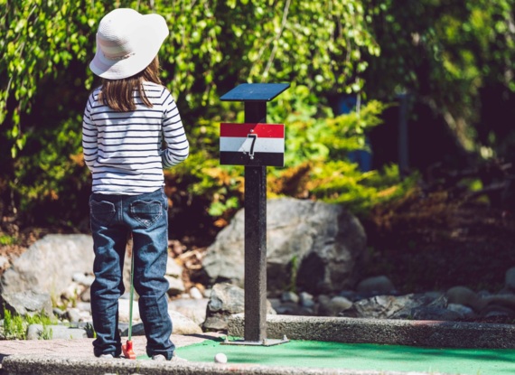 Kind spielt Minigolf auf grünem Platz mit nummeriertem Loch vor natürlicher Felsen- und Blattkulisse