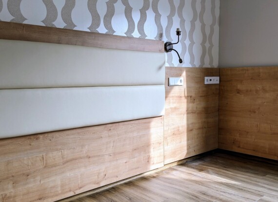Modern hotel room corner with wood panel walls, padded headboard, subtle patterned wallpaper, and natural light on wooden floor