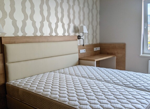 Modern hotel room with a queen bed featuring a padded headboard, wall-mounted lamps, wooden desk, and city view through window