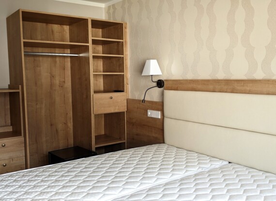 Hotel room with twin mattresses, upholstered headboard, wood wardrobe with shelves and drawer, soft cream wallpaper, and wall-mounted lamp