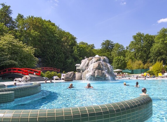 Beheizter Außenpool mit geschwungenen Fliesenrändern, roter Bogenbrücke, felsigem Wasserfall, umgeben von grünen Bäumen und sonnenbadenden Gästen
