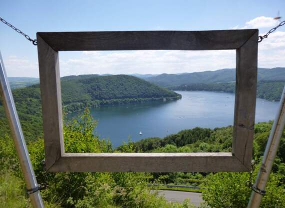 Holzrahmen an Ketten über einem ruhigen See mit grünen Hügeln und klarem Himmel