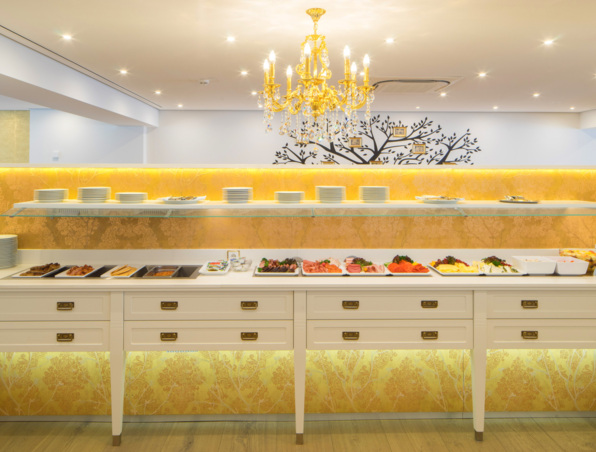 Elegantes Frühstücksbuffet mit verschiedenen Käsesorten, Aufschnitt, Früchten und Tellern unter einem goldenen Kronleuchter in hellem Speiseraum