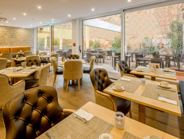Helles Hotelrestaurant mit bequemen Polsterstühlen, Holztischen zum Frühstück und Terrasse mit Sitzgelegenheiten an Steinwand