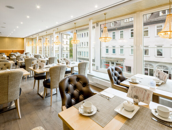 Helles Hotelrestaurant mit großen Fenstern, eleganten Kronleuchtern, gepolsterten Stühlen und liebevoll gedeckten Frühstückstischen.