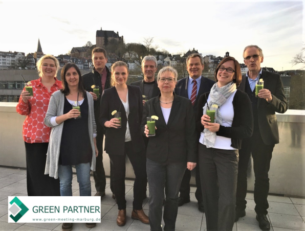 Neun Personen genießen grüne Smoothies auf einer Hoteldachterrasse mit Blick auf historische Stadt und Schloss bei Sonnenuntergang