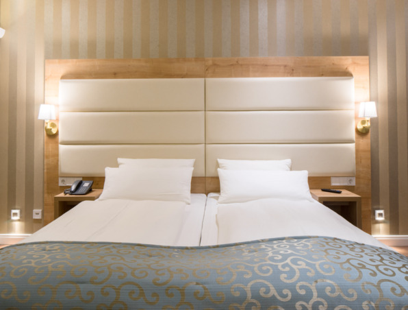 Modernes Hotelzimmer mit zwei Einzelbetten, gepolstertem weißem Kopfteil, beige gestreifter Tapete, Nachttischlampen und blauem Bettüberwurf mit Muster