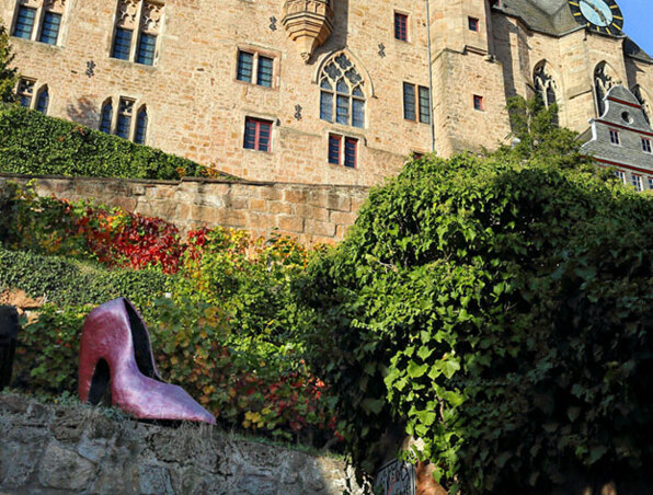 Historisches Steingebäude mit gotischen Fenstern über üppigem grünem Efeu und großer roter High-Heel-Skulptur im Freien
