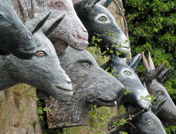 Skulptierte Tierköpfe, darunter Ziegen und Wildschwein, an einer Steinmauer mit grüner Vegetation